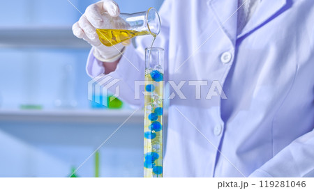 Close up shot from front view, yellow liquid from a glass erlenmeyer flask being poured into a measuring cylinder filled by clear water and blue hydrogel. Copy space for text Close up shot from front view, yellow liquid from a glass erlenmeyer flask being poured into a measuring cylinder filled by clear water and blue hydrogel. Copy space for text 119281046