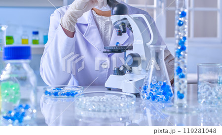 The experiment scene with blue and white hydrogel, a scientist is sitting at the lab table and looking into microscope to observe a blue gel ball. Front view, minimal laboratory background The experiment scene with blue and white hydrogel, a scientist is sitting at the lab table and looking into microscope to observe a blue gel ball. Front view, minimal laboratory background 119281048