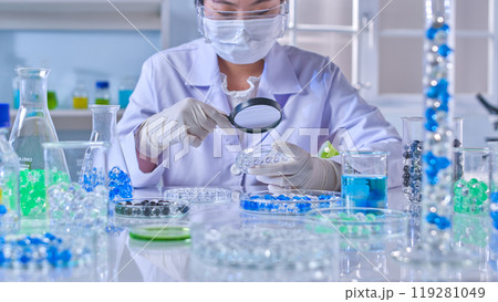 Advertising photo of experimental theme, a professional scientist sitting at the lab table, which covered by various laboratory glassware contain colorful gel ball, observed by magnifying glass 119281049