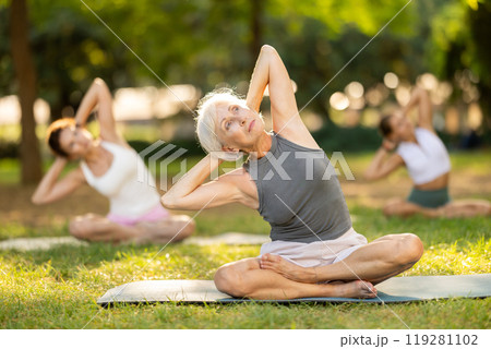 Happy old lady and class of calm sports women in activewear stretching muscles and doing tilts while sitting in lotus pose on mats during yoga training together at city park Happy old lady and class of calm sports women in activewear stretching muscles and doing tilts while sitting in lotus pose on mats during yoga training together at city park 119281102