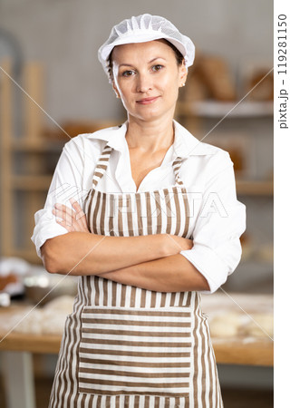 Confident young female bakery in apron posing with arms crossed indoors in bakery kitchen 119281150