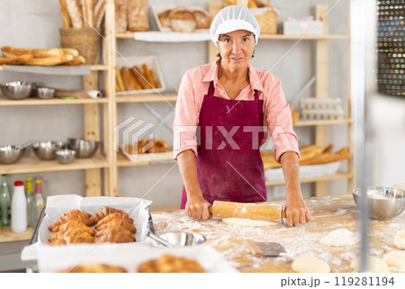 Portrait of happy mature female professional baker in uniform standing and posing in bakery during working day 119281194