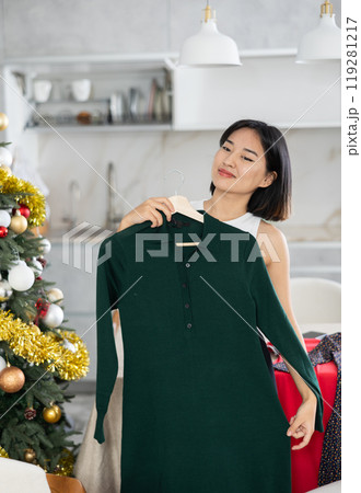 Happy young Asian girl trying to choose a dress standing next to Xmas-tree 119281217