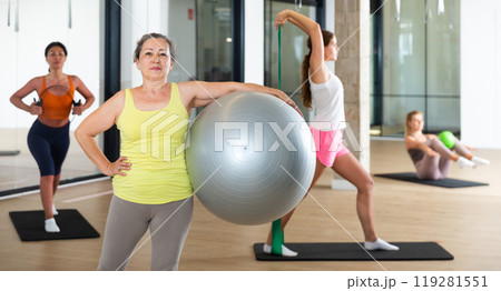 Portrait of mature woman standing with fitness ball during group training at gym Portrait of mature woman standing with fitness ball during group training at gym 119281551