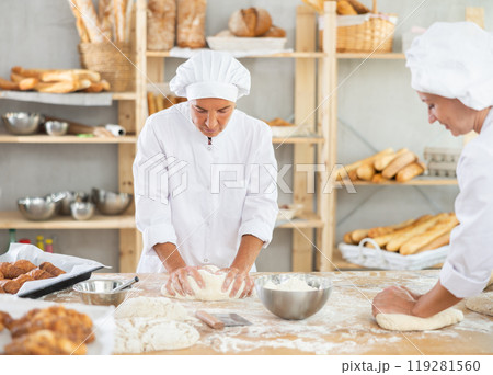 Professional mature female baker with assistant kneading dough and portioning dough in pieces during daily work in bakery Professional mature female baker with assistant kneading dough and portioning dough in pieces during daily work in bakery 119281560