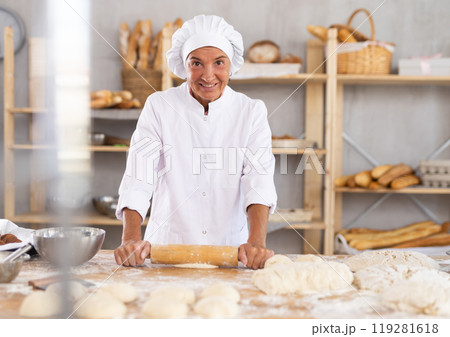Smiling mature female baker rolling out dough in bakery 119281618