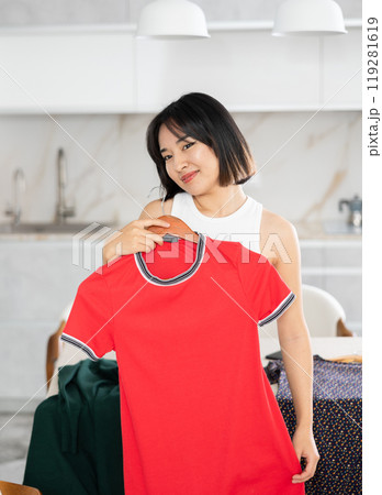 Happy young Asian girl trying to choose a dress in the kitchen 119281619
