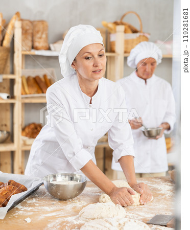 Skilled smiling young female professional baker in white uniform standing at work table and kneading dough during working day in bakery Skilled smiling young female professional baker in white uniform standing at work table and kneading dough during working day in bakery 119281681