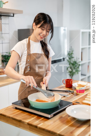 A beautiful, happy Asian woman in an apron is enjoying cooking breakfast in the kitchen. A beautiful, happy Asian woman in an apron is enjoying cooking breakfast in the kitchen. 119282026