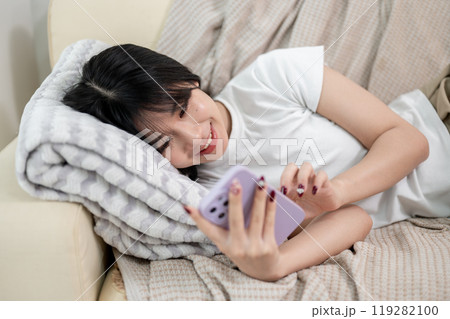 A cheerful Asian woman is enjoying reading chats on her smartphone while lying on a couch. A cheerful Asian woman is enjoying reading chats on her smartphone while lying on a couch. 119282100