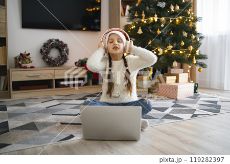 Little girl enjoys music on laptop near Christmas tree Little girl enjoys music on laptop near Christmas tree 119282397
