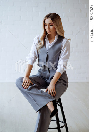 Young blonde woman in gray vest and trousers sits on black chair on white background.  119282525