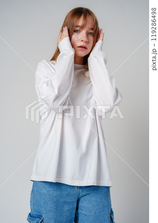 A young woman with long hair hides her face in a white sweater against a plain background 119286498
