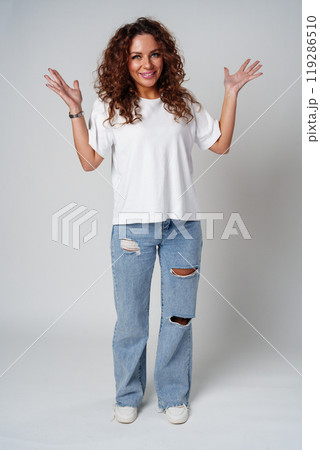 Cheerful woman with curly hair poses with open arms in a white shirt against a plain background 119286510