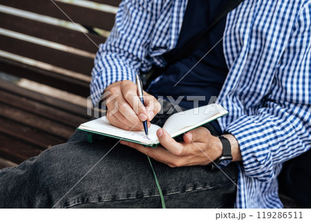 A person writes in a notebook while sitting on a park bench A person writes in a notebook while sitting on a park bench 119286511