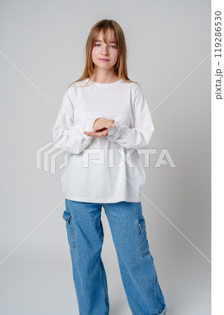 Casual indoor portrait of a young woman in a white sweater and blue jeans posing with her hands together 119286530