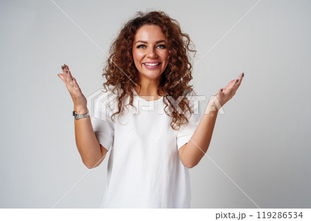Cheerful woman with curly hair poses with open arms in a white shirt against a plain background Cheerful woman with curly hair poses with open arms in a white shirt against a plain background 119286534