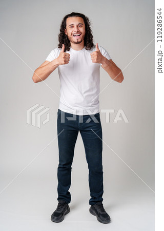 Young man with curly hair gives a thumbs-up in a casual outfit against a neutral background 119286544