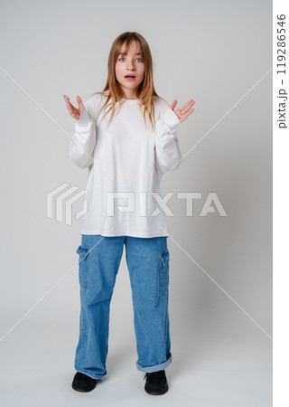 A surprised child wearing a white long-sleeve shirt and blue jeans expresses confusion in a studio setting 119286546