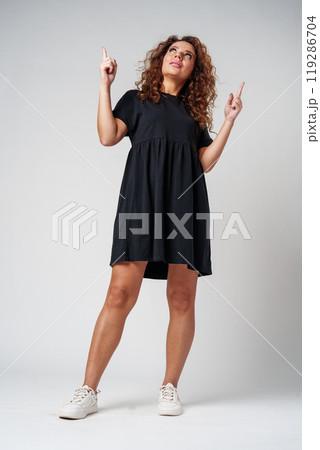 Cheerful young woman with curly hair giving a thumbs up while smiling against a neutral background 119286704