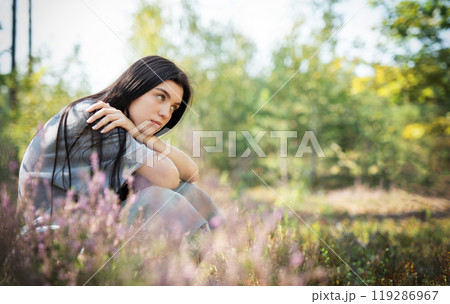 A young woman sitting among wildflowers in a serene forest, lost in thought during a sunny afternoon 119286967