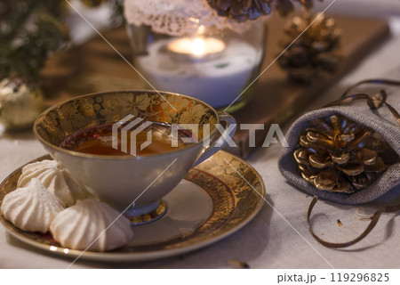 Winter still life. Cup of tea with meringues on a cozy table with a linen tablecloth on it. Beside a candle and decorative pine cones in a bag. Cozy atmosphere. Porcelain cup with gilding and painting Winter still life. Cup of tea with meringues on a cozy table with a linen tablecloth on it. Beside a candle and decorative pine cones in a bag. Cozy atmosphere. Porcelain cup with gilding and painting 119296825