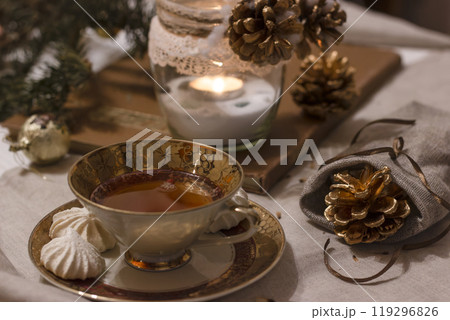 Cozy winter evening tea setting with cookies, pinecones, and candlelight at home. Cup of tea with cookies . Decorative pine cones on the linen tablecloth. Candle enhances the cozy atmosphere. Cozy winter evening tea setting with cookies, pinecones, and candlelight at home. Cup of tea with cookies . Decorative pine cones on the linen tablecloth. Candle enhances the cozy atmosphere. 119296826