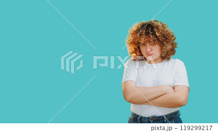 Offended expression. Grumpy face. Body shaming. Frowning upset overweight shy chubby girl with crossed arms isolated on blue free space background. 119299217