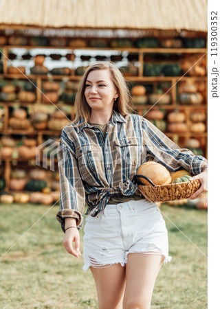 Young woman holding a basket of pumpkins near a haystack on a farm field. Outdoor autumn harvest scene. Fall season and agriculture concept for design and print 119300352