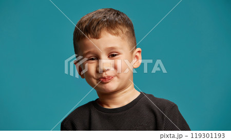 Boy exudes playful and cheeky charm, with slight smile, looking like he's trying to hold back r secret against blue studio background. 119301193