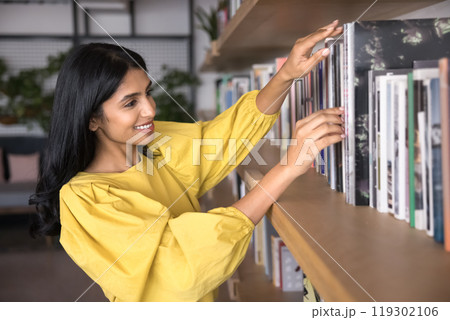 Happy young Indian reading fan woman picking book from bookshelf Happy young Indian reading fan woman picking book from bookshelf 119302106
