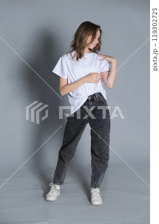 A young girl stands in a relaxed pose, adjusting her t-shirt while looking down. Her casual style, with jeans and sneakers, reflects a laid-back and modern sense of fashion. A young girl stands in a relaxed pose, adjusting her t-shirt while looking down. Her casual style, with jeans and sneakers, reflects a laid-back and modern sense of fashion. 119302725