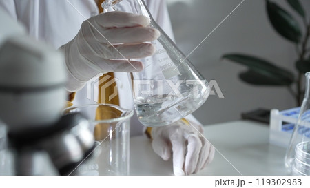Unknown scientist wearing white protective gloves is carefully shaking up a clear chemical solution inside an Erlenmeyer flask in a laboratory, close up. Science and medicine 119302983