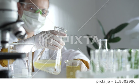 Woman scientist wearing a lab coat, white gloves, and a mask is pouring a yellow oily liquid from one beaker to another in a laboratory setting, close up. Science and medicine Woman scientist wearing a lab coat, white gloves, and a mask is pouring a yellow oily liquid from one beaker to another in a laboratory setting, close up. Science and medicine 119302986