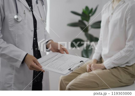 Doctor and a patient discussing something. The female physician, wearing a white medical coat is keeping clipboard with a medical notes besides a young woman during a consultation in the clinic Doctor and a patient discussing something. The female physician, wearing a white medical coat is keeping clipboard with a medical notes besides a young woman during a consultation in the clinic 119303000