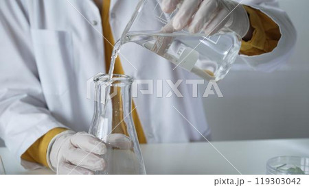 Unknown scientist with a lab coat and white protective gloves is carefully pouring a liquid from a beaker into an Erlenmeyer flask, close up. Science and medicine concept 119303042