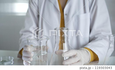 A scientist with a lab coat and white protective gloves is carefully pouring a liquid from one beaker to another, close up. Science and medicine concept A scientist with a lab coat and white protective gloves is carefully pouring a liquid from one beaker to another, close up. Science and medicine concept 119303043
