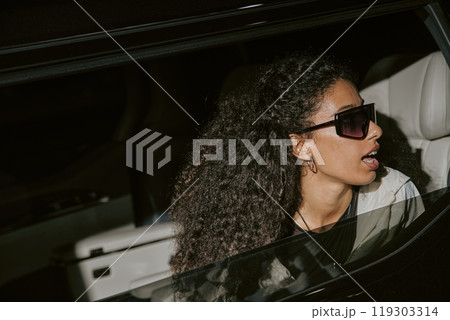 Shot through opened car window of glamorous Middle Eastern female celebrity in futuristic sunglasses caught unawares by paparazzi sitting in back seat of limousine, camera flash, copy space 119303314