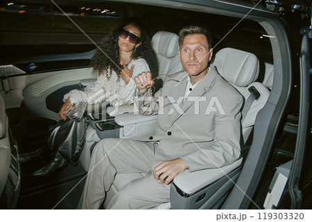 Stylish male celebrity in classy grey suit surprised by paparazzis camera flash sitting in back seat of car with fashionable curly woman in silver outfit, while riding in night city Stylish male celebrity in classy grey suit surprised by paparazzis camera flash sitting in back seat of car with fashionable curly woman in silver outfit, while riding in night city 119303320