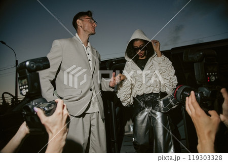 Low angle shot of glamorous female Middle Eastern celebrity pulling up hood for privacy caught by paparazzi stepping out of car, accompanied by well dressed man in suit at night street in city 119303328