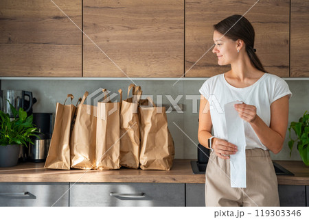 Young woman with receipt from the store next to paper bags full of food in kitchen. Groceries ordered online and delivered by courier 119303346
