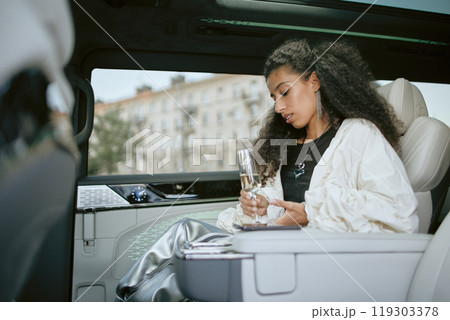 Side view of glamorous Middle Eastern woman wearing elegant silver jewellery sitting in back seat of car while holding glass of champagne embodying celebrity lifestyle, copy space Side view of glamorous Middle Eastern woman wearing elegant silver jewellery sitting in back seat of car while holding glass of champagne embodying celebrity lifestyle, copy space 119303378