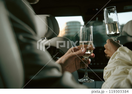 Close up on hands of unrecognizable couple holding champagne flutes clinking glasses while celebrating anniversary in back seat of car, copy space Close up on hands of unrecognizable couple holding champagne flutes clinking glasses while celebrating anniversary in back seat of car, copy space 119303384