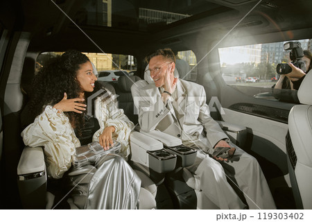 Medium shot of smiling male celebrity in classy suit talking to young Middle Eastern girlfriend sitting in back seat of car, while enjoying paparazzis attention living in public eye 119303402