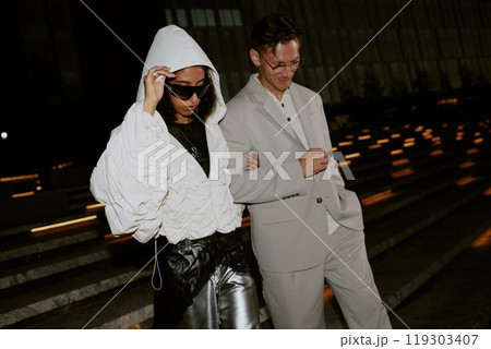 Famous female Middle Eastern celebrity wearing sunglasses and trendy jacket pulling up hood to hide from paparazzis attention while walking with male partner in city street at night time, copy space Famous female Middle Eastern celebrity wearing sunglasses and trendy jacket pulling up hood to hide from paparazzis attention while walking with male partner in city street at night time, copy space 119303407