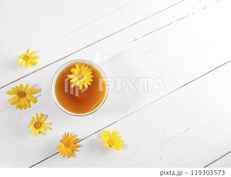 Top view one white cup of calendula tea and flower heads around it on white wooden table. Copy space 119303573