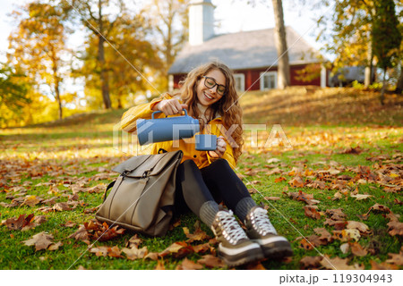Woman traveler in yellow coat sits in clearing among yellow leaves and drinks hot drink from thermos Woman traveler in yellow coat sits in clearing among yellow leaves and drinks hot drink from thermos 119304943