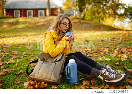 Woman traveler in yellow coat sits in clearing among yellow leaves and drinks hot drink from thermos Woman traveler in yellow coat sits in clearing among yellow leaves and drinks hot drink from thermos 119304945