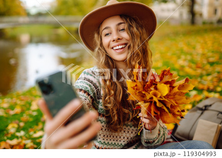 Selfie time. Beautiful woman takes selfie on smartphone in hat on meadow in autumn park on plaid. 119304993