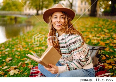 Beautiful curly woman in hat on mat with book in autumn park. Relaxation, solitude with nature. Beautiful curly woman in hat on mat with book in autumn park. Relaxation, solitude with nature. 119304995